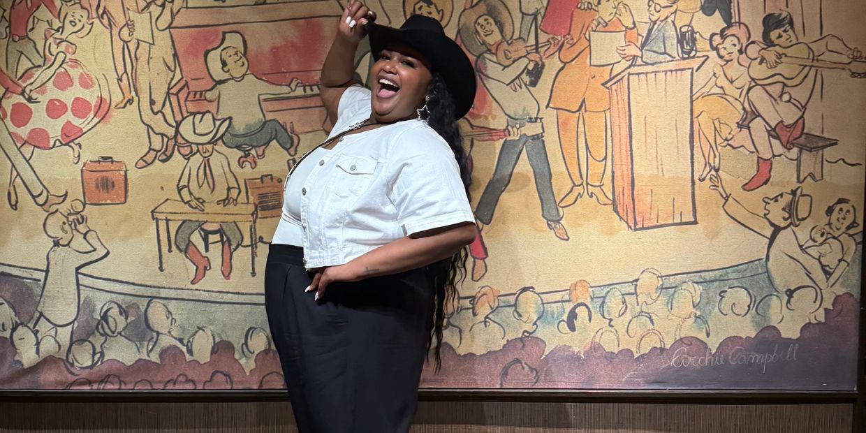A joyful woman in a cowboy hat poses in front of a colorful Grand Ole Opry mural.