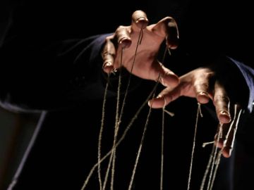 Hands manipulating puppet strings in dark lighting.