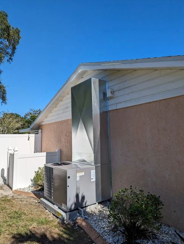 Outdoor HVAC unit with metal ductwork attached to a house under clear blue sky.