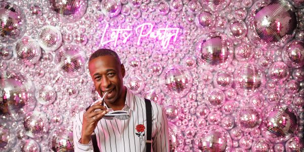 Man poses with sunglasses in front of shiny disco ball wall.