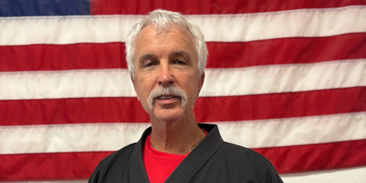 Man with white hair and mustache in martial arts uniform with American flag backdrop.