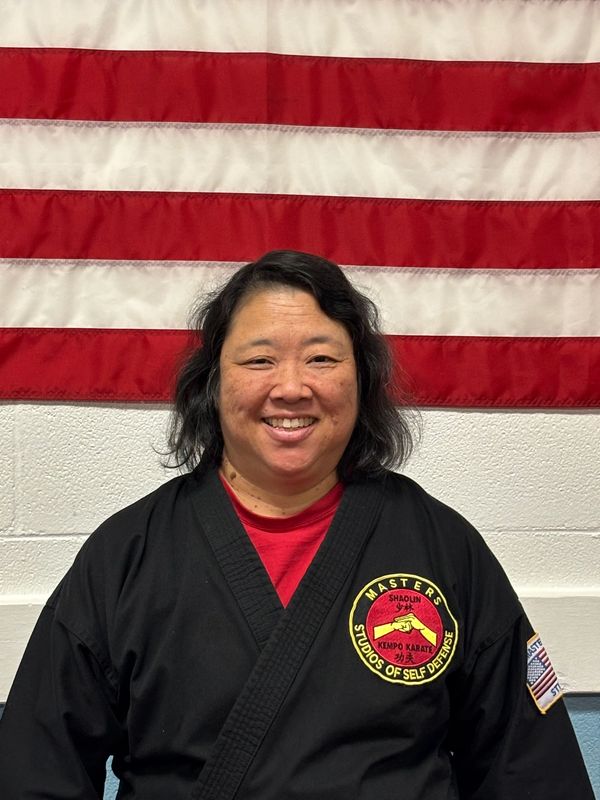 A smiling martial arts master in black uniform stands in front of an American flag.
