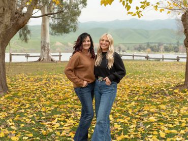 Two women posing outdoors among fallen leaves.