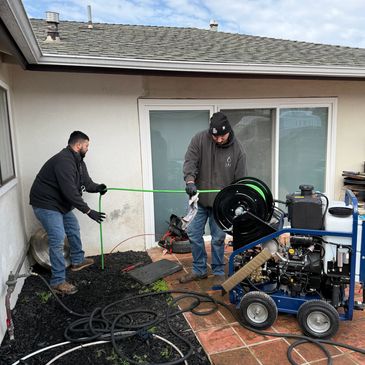 Two men working with a hose and equipment outside a house.