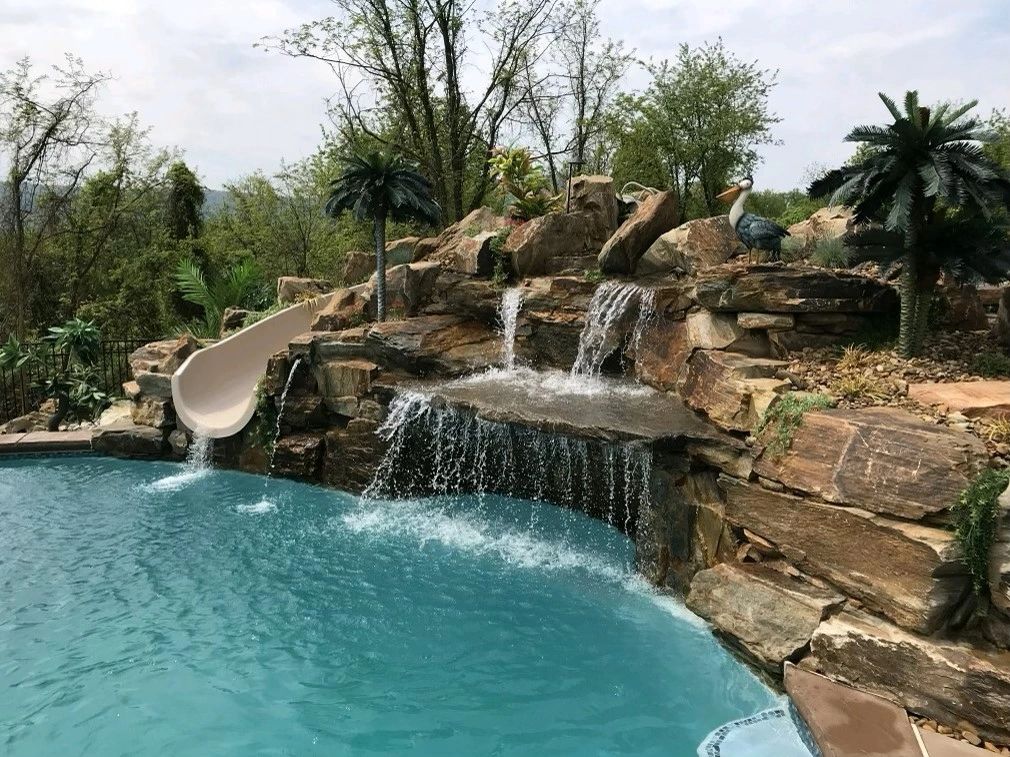 Two-tier boulder waterfall with waterslide, cave, and plants. Riverstone, palm trees, and mountains.