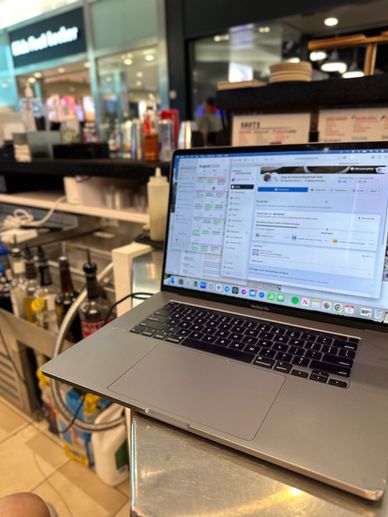 Laptop open on bar counter with bottles and supplies in background.