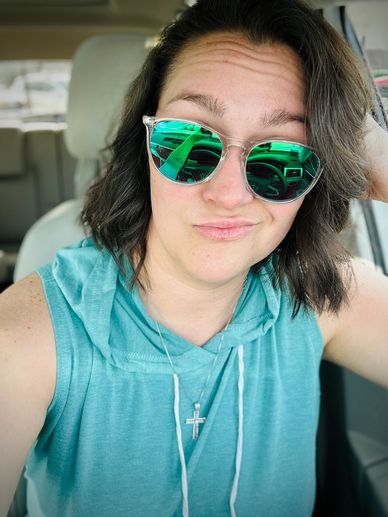 Woman in teal sleeveless hoodie wearing green reflective sunglasses with a smirk.