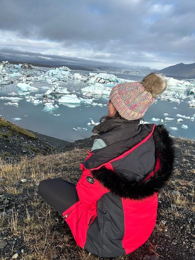 Stephanie in red coat and knit hat sitting by an iceberg lagoon.