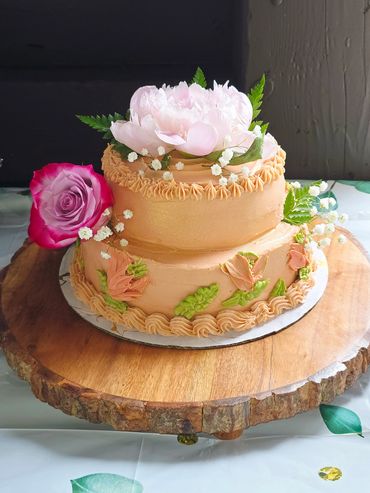 Two-tier peach cake adorned with fresh flowers on a wooden slab.