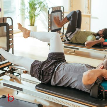 Two men exercising on Pilates reformer machines in a bright studio.