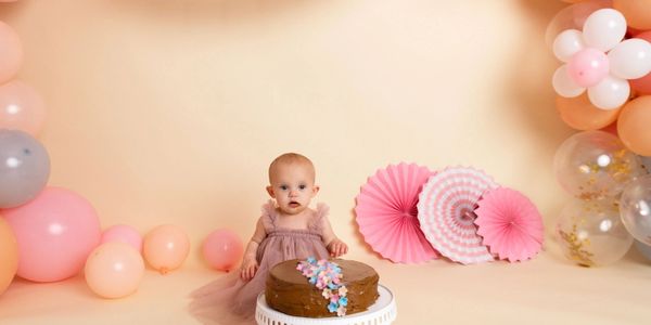 A baby girl in a pink dress with a chocolate cake and pastel balloons.