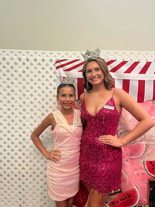 Two young beauty queens in sparkling dresses and crowns smile together.