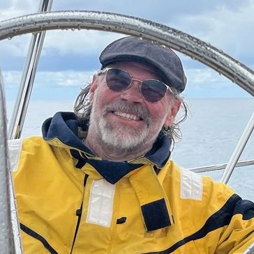 A man in a yellow jacket and cap smiles while steering a boat.