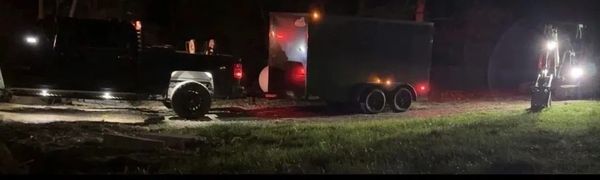 A dark truck with a trailer and a lit excavator at night on a dirt road.