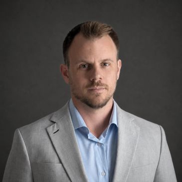 Portrait of a man in a light gray suit jacket against a dark background.