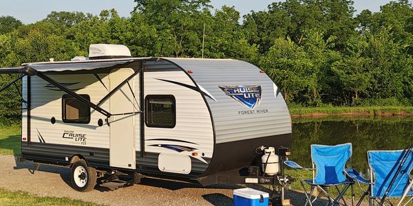 Travel trailer parked near a lake with camping chairs and cooler.