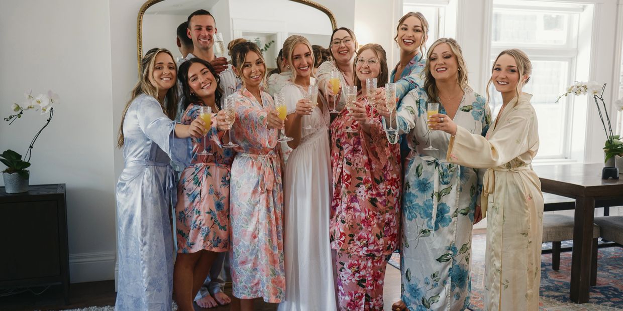 Group of bridesmaids and bride in robes toasting with drinks indoors.