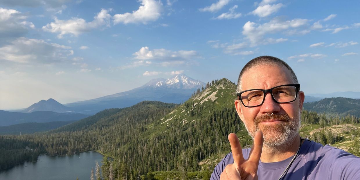 Man showing peace sign with mountain and lake backdrop.