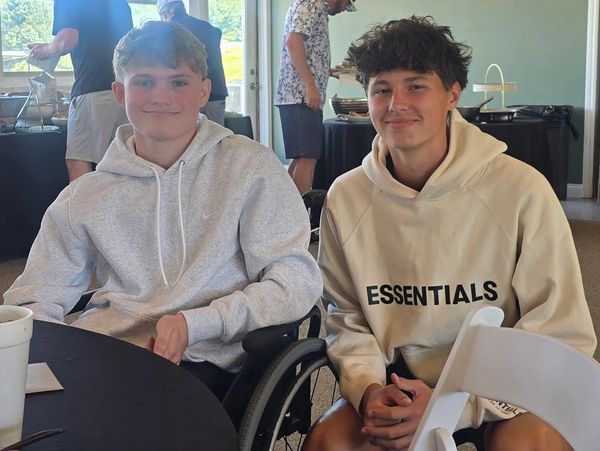 Two young men sitting together, one in a wheelchair, both wearing hoodies indoors.