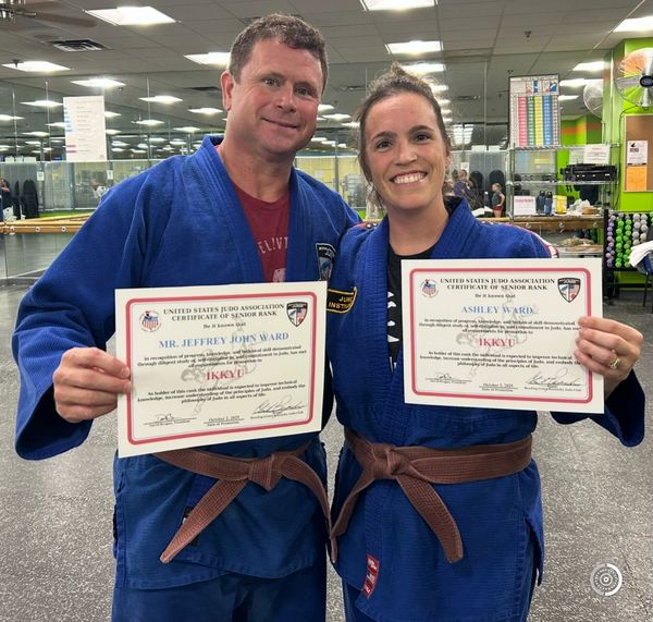 Two people in judo uniforms proudly holding senior rank certificates.