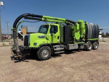 Bright green industrial vacuum truck parked on a dirt lot under clear blue sky.