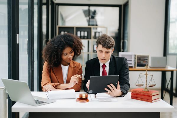 Two professionals discuss a tablet at a modern office desk, with a gavel and law books in view