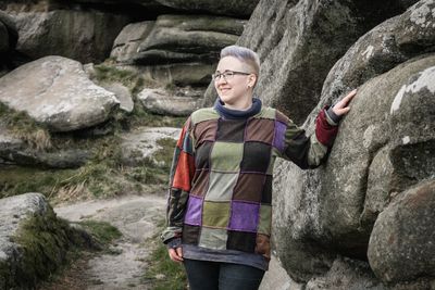 A photo of Tori standing among large rocks in the Peak District, looking to their right and smiling.