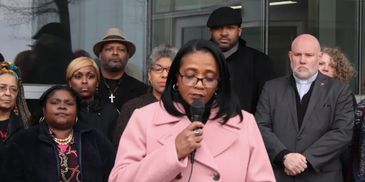 Woman in pink coat speaking into microphone with attentive group behind her.