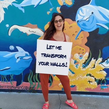 Artist standing in front of full color mural holding a sign that says "Let me transform your walls!"