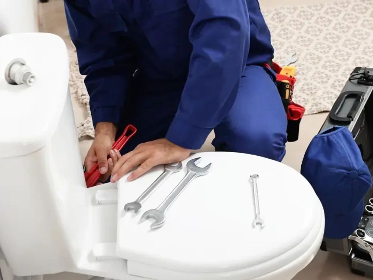 A plumber fixing a toilet with tools on the seat and a toolbox nearby.