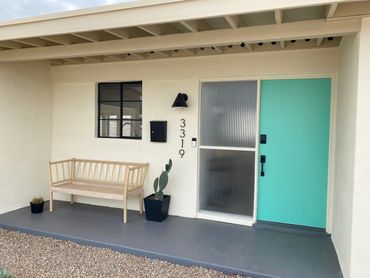 Minimalist porch with a turquoise door, light wooden bench, and potted cacti.