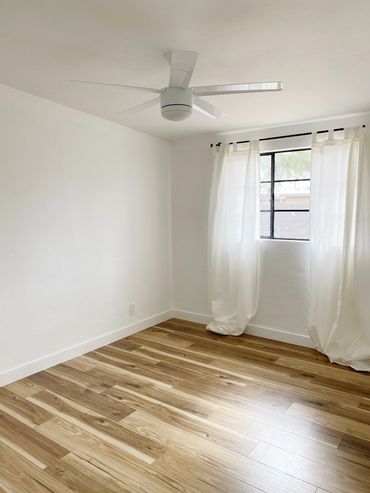 Empty room with wooden floor, white walls, ceiling fan, and window with sheer curtains.