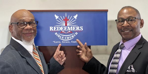 Two men in suits pointing at a Redeemers Bible Church sign.