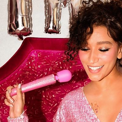 A woman wearing pink sparkles holding a massager with pink sparkles like a microphone while smiling.