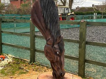 A wooden horse sculpture with its head lowered, outdoors near a fence.