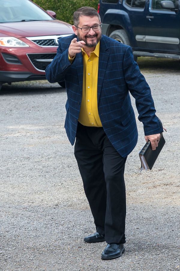 Smiling man in a blue blazer pointing and holding a black folder outdoors.