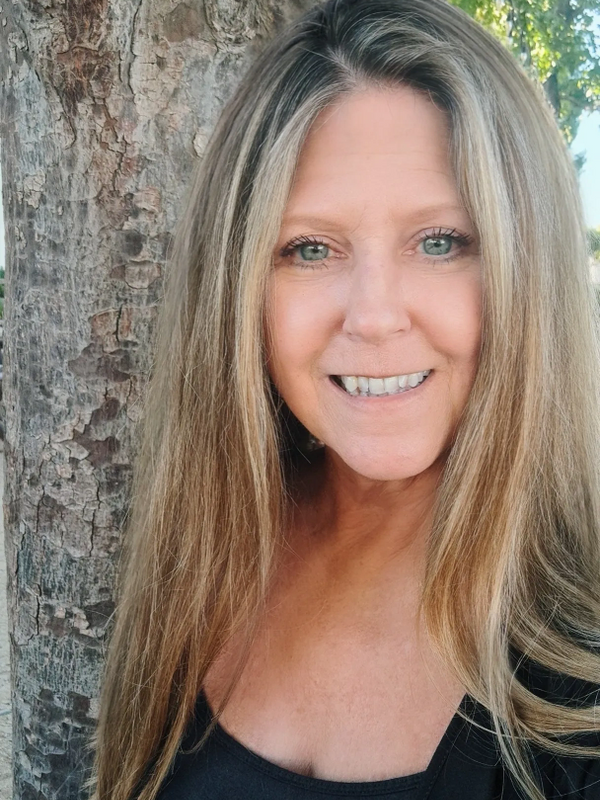 Smiling woman with long blonde hair standing by a tree outdoors.