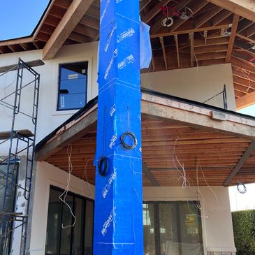 Under-construction house with blue-wrapped pillar and scaffolding on a sunny day.