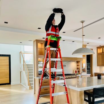 Worker on a ladder fixing a ceiling fixture in a modern kitchen.