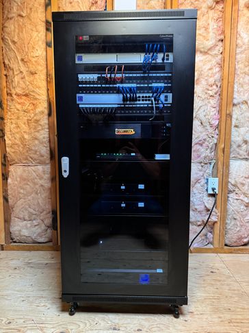 Black server rack with cables and security equipment in an unfinished room.