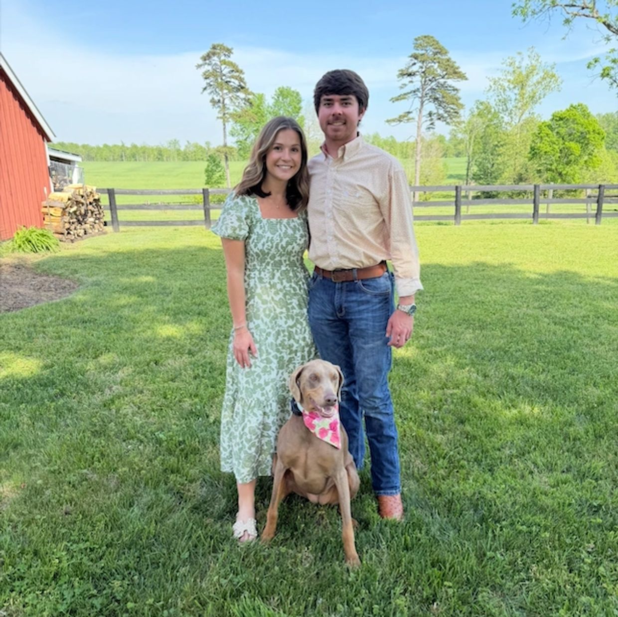 A smiling couple stands with their dog in a sunny green backyard.