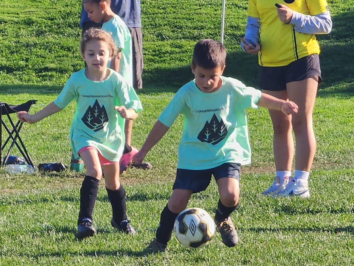 Two children playing soccer on a grassy field with a coach nearby.