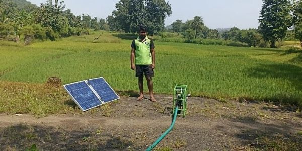 Man stands near a solar panel and irrigation pump in a green field.