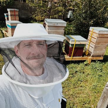 Beekeeper smiling in protective suit near beehives outdoors.