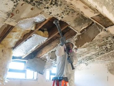 Worker in protective gear removing moldy ceiling drywall.