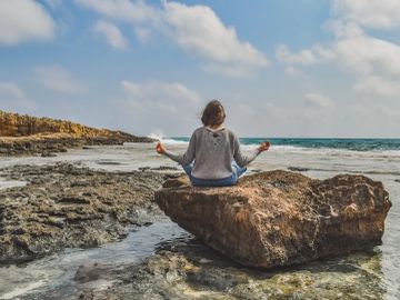 Person meditating on a large rock by the sea under a partly cloudy sky.