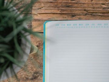 Open lined notebook with months header on wooden table beside plant.
