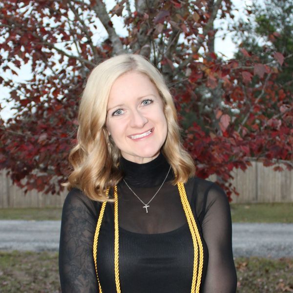 Smiling woman with blonde hair in a black outfit and gold honor cords outdoors.