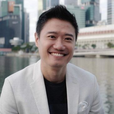 Smiling man in white blazer by a city waterfront with skyscrapers.