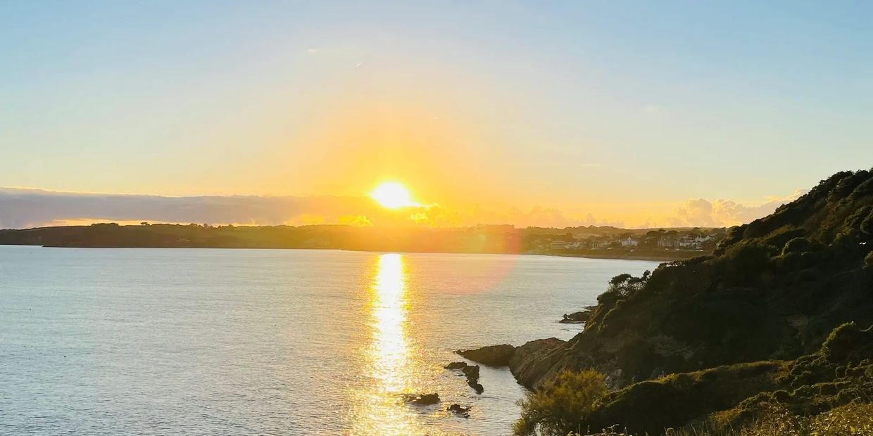 Sunset over a calm coastal bay with hills and vegetation.